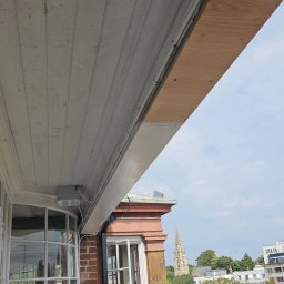 DECOLUX - Widok z balkonu na miasto, fragment zniszczonego sufitu z odpadającą farbą i nowa, surowa deska montowana do sufitu balkonu.