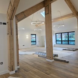 DECOLUX - Przestronne wnętrze w trakcie remontu z drewnianymi belkami stropowymi, zabezpieczona podłoga, drabina i nowoczesne okrągłe lampy sufitowe.