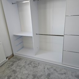 DECOLUX - Wnętrze szafy wnękowej z przesuwnymi drzwiami, widoczne półki i drążki na ubrania, szara wykładzina dywanowa na podłodze, grzejnik na ścianie obok.