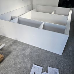 DECOLUX - Biała, modułowa konstrukcja regału z podziałem na kwadratowe półki, częściowo zamontowana, z instrukcją montażu rozłożoną na szarym dywanie w tle białej ściany z gniazdkiem elektrycznym.