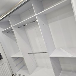 DECOLUX - Wnętrze szafy wnękowej z białymi półkami i drążkami na ubrania, widoczne numerki na elementach konstrukcyjnych, fragment okna z zasłoną i grzejnik w tle.