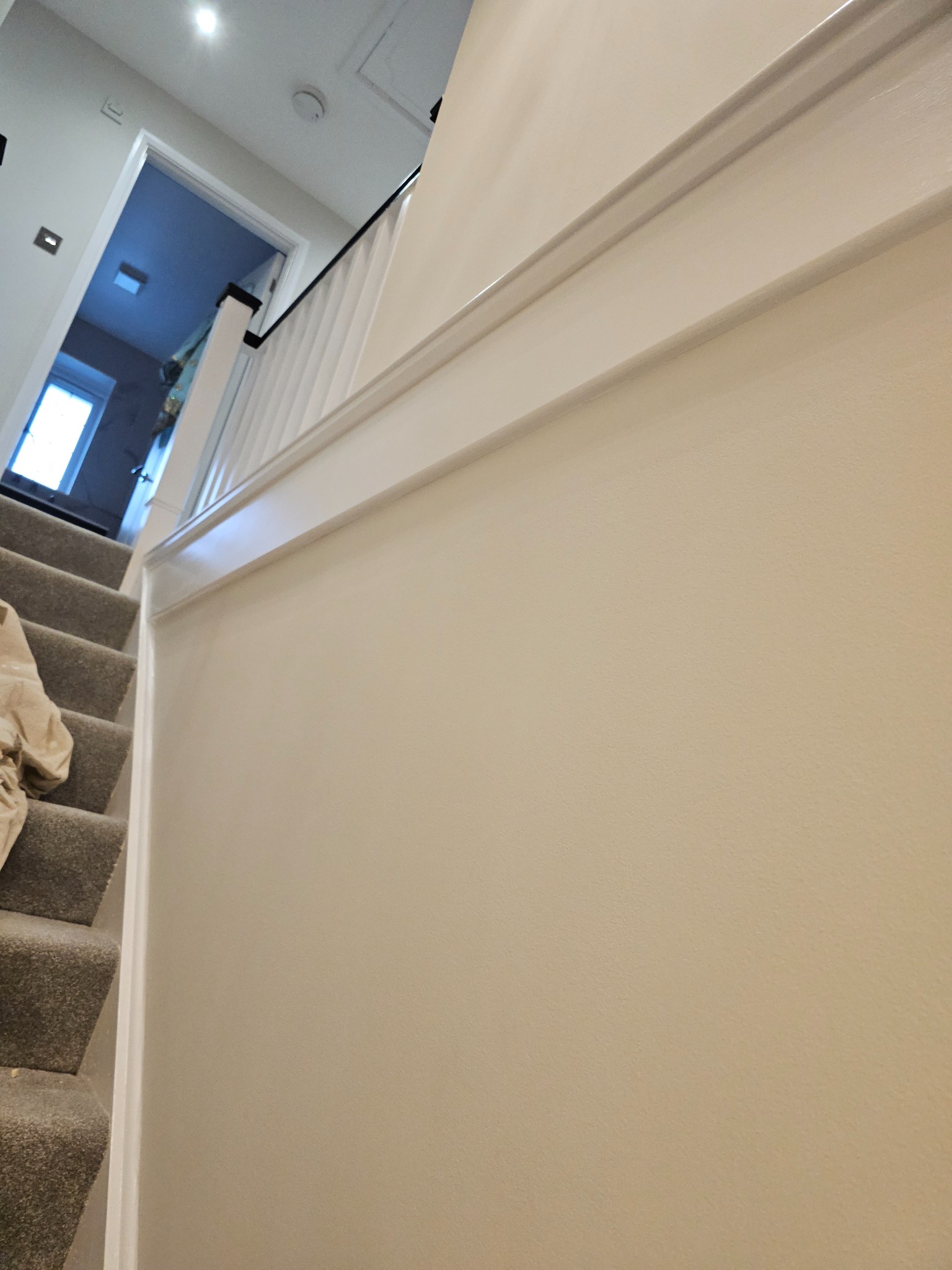 Świeżo pomalowana ściana klatki schodowej w odcieniu beżu z białą listwą dekoracyjną, widoczne schody pokryte wykładziną dywanową w odcieniach szarości.