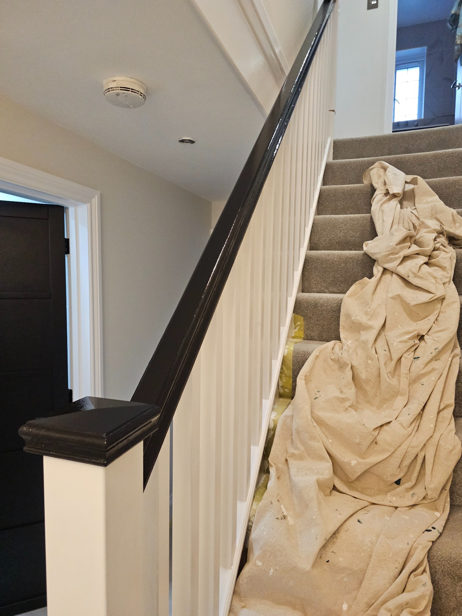 Schody zabezpieczone płachtą malarską, świeżo pomalowana, błyszcząca, czarna balustrada kontrastuje z białymi tralkami i szarą wykładziną schodową.