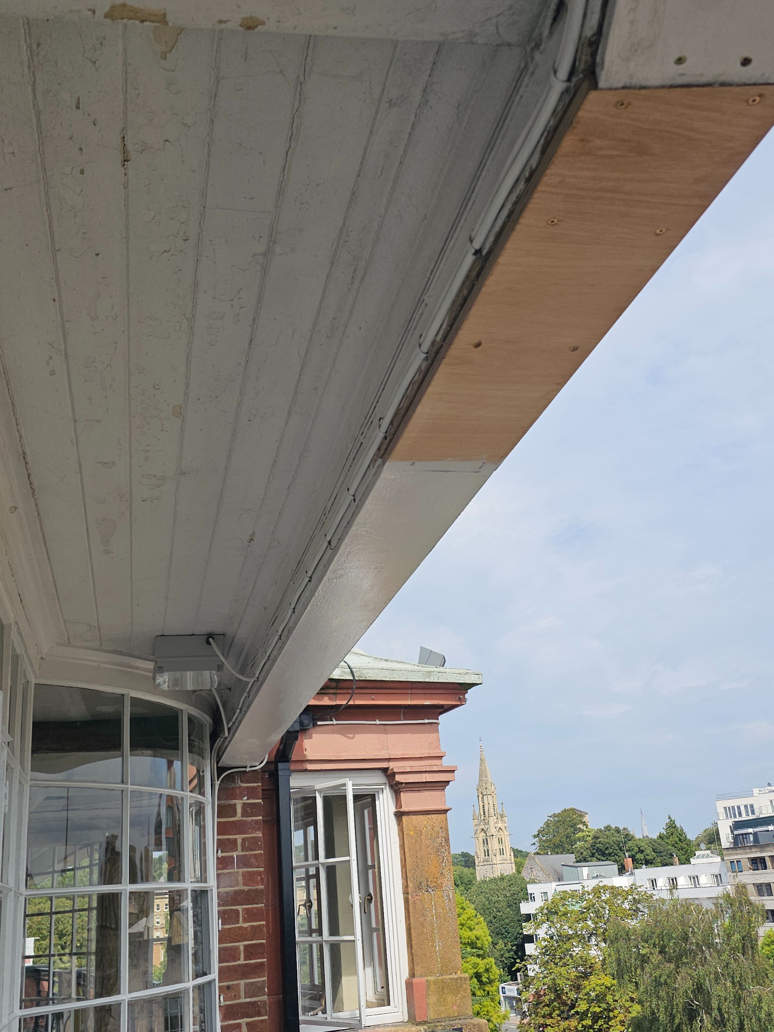 Widok z balkonu na miasto, fragment zniszczonego sufitu z odpadającą farbą i nowa, surowa deska montowana do sufitu balkonu.