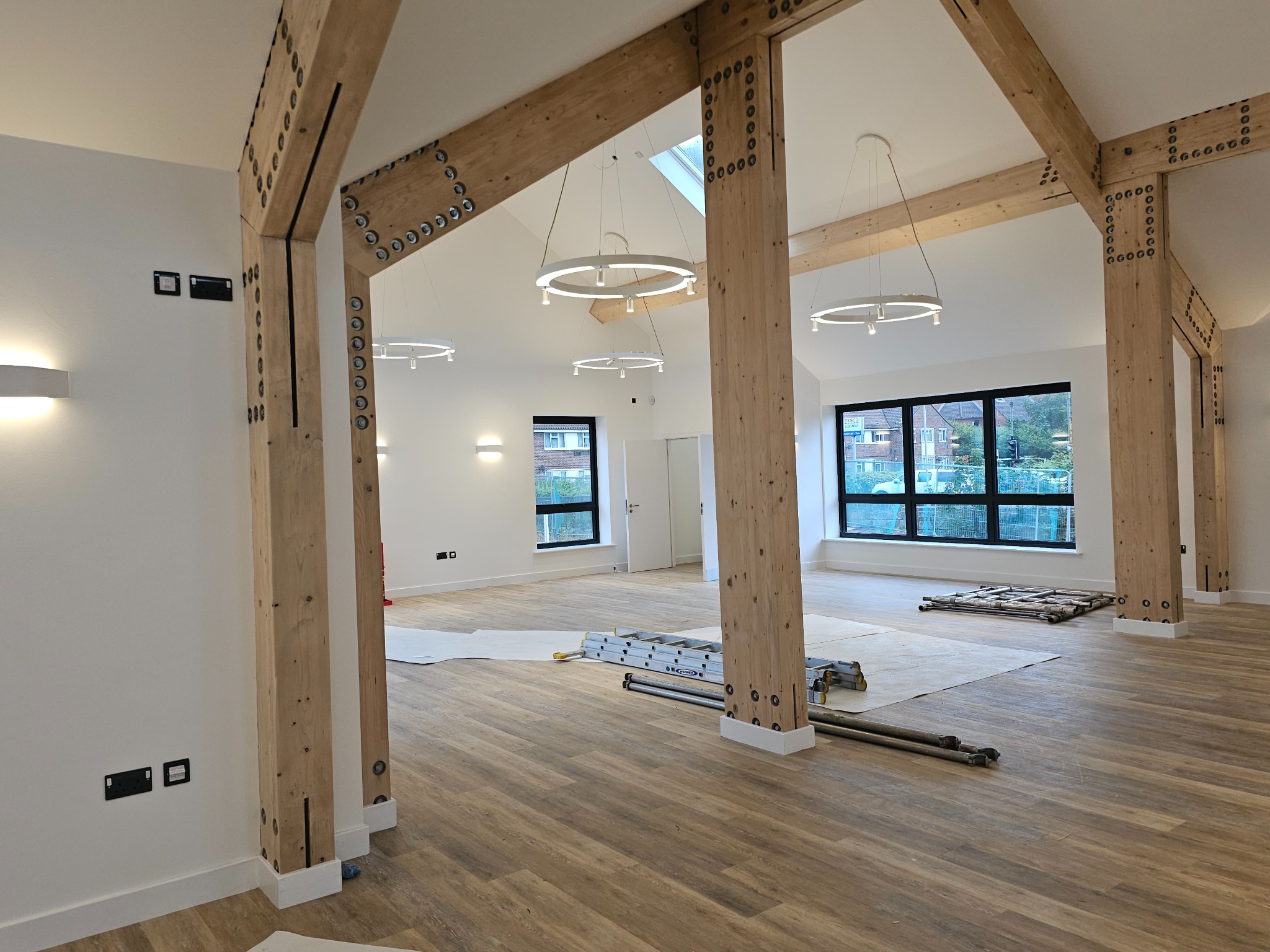 Przestronne wnętrze w trakcie remontu z drewnianymi belkami stropowymi, zabezpieczona podłoga, drabina i nowoczesne okrągłe lampy sufitowe.