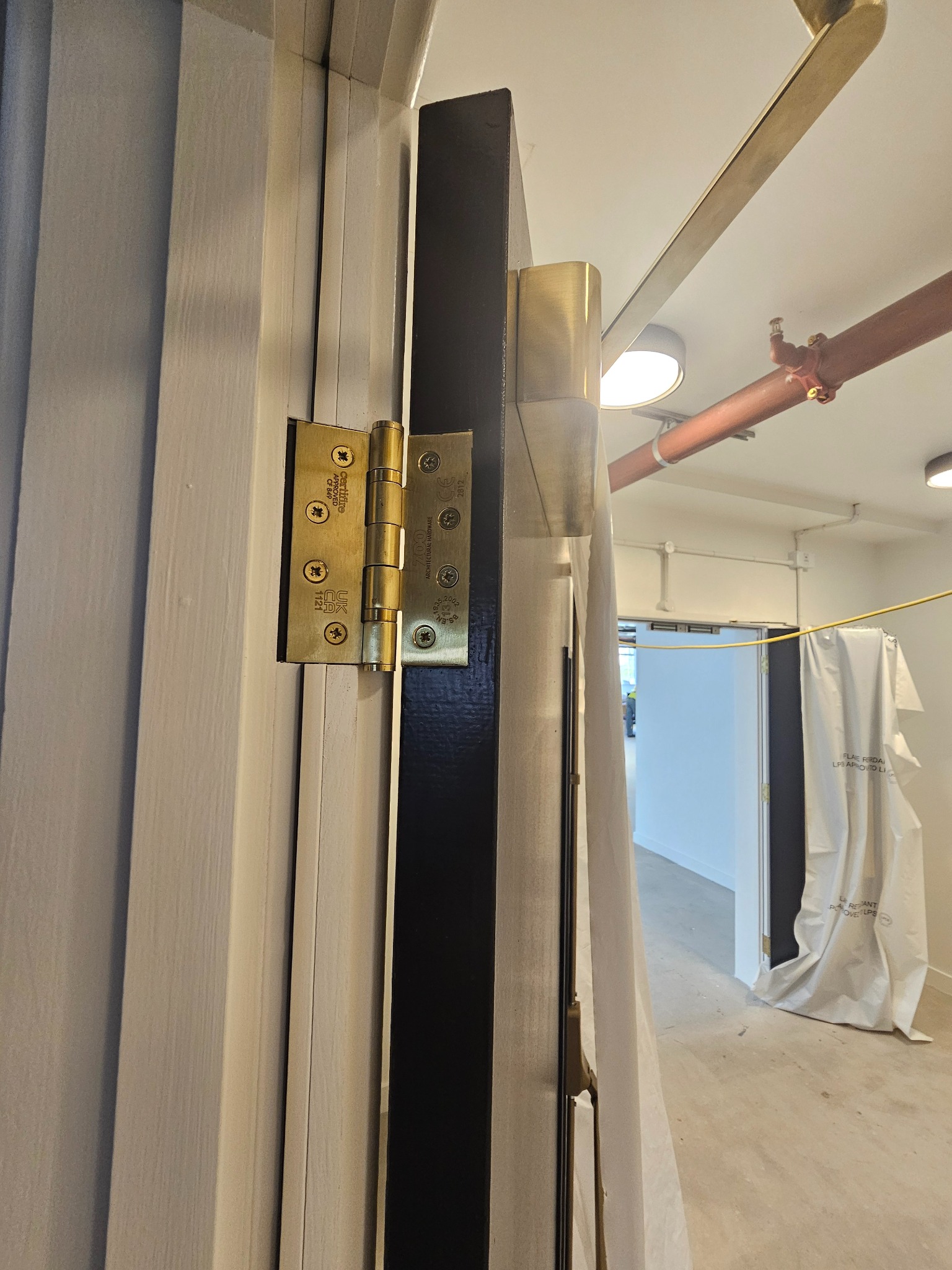 Detal złote zawiasy na świeżo pomalowanych drzwiach w odcieniach bieli i czerni, widoczne zabezpieczenia podłogi i rur na suficie podczas remontu.