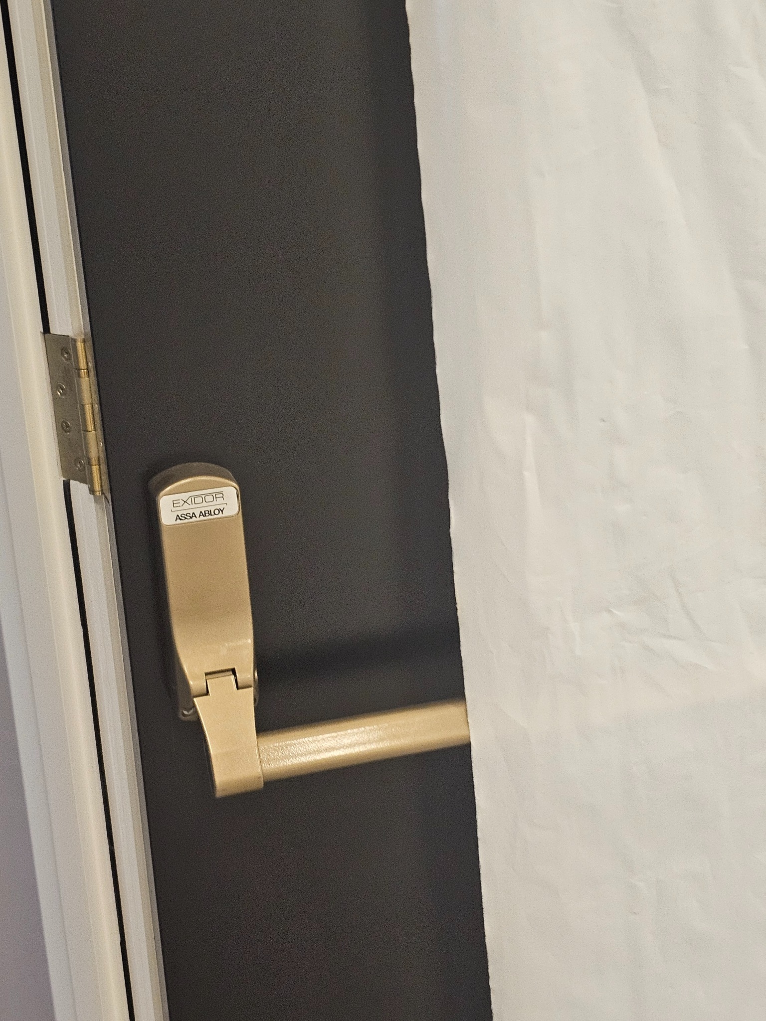 Szara ściana z zamontowaną, złotą klamką antypaniczną marki Exidor, częściowo zasłonięta białą folią ochronną.