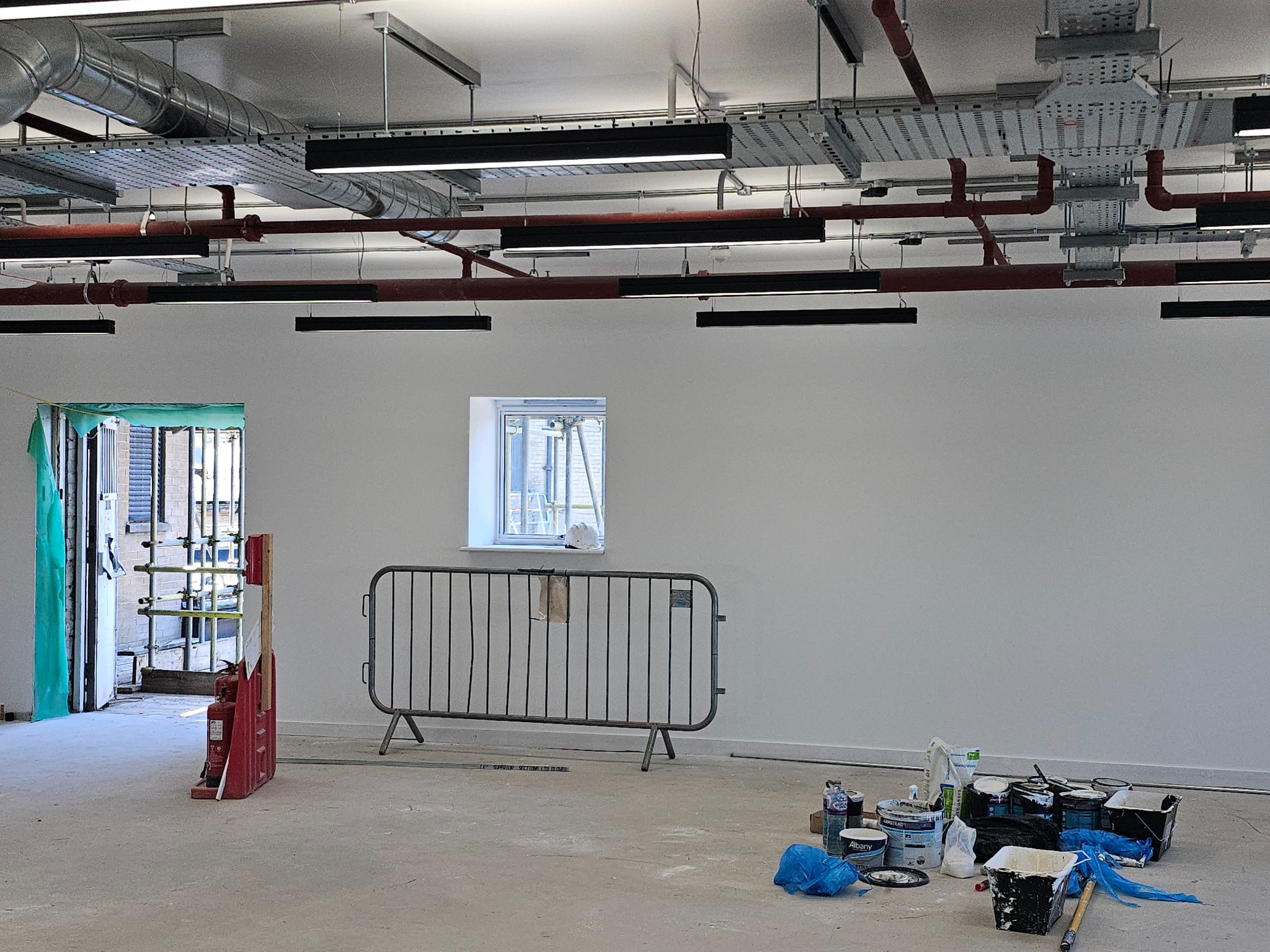 Wnętrze w trakcie prac wykończeniowych: ściany pomalowane na biało, widoczne puszki z farbą, zabezpieczenie budowlane, instalacje pod sufitem, okno z rusztowaniem na zewnątrz.