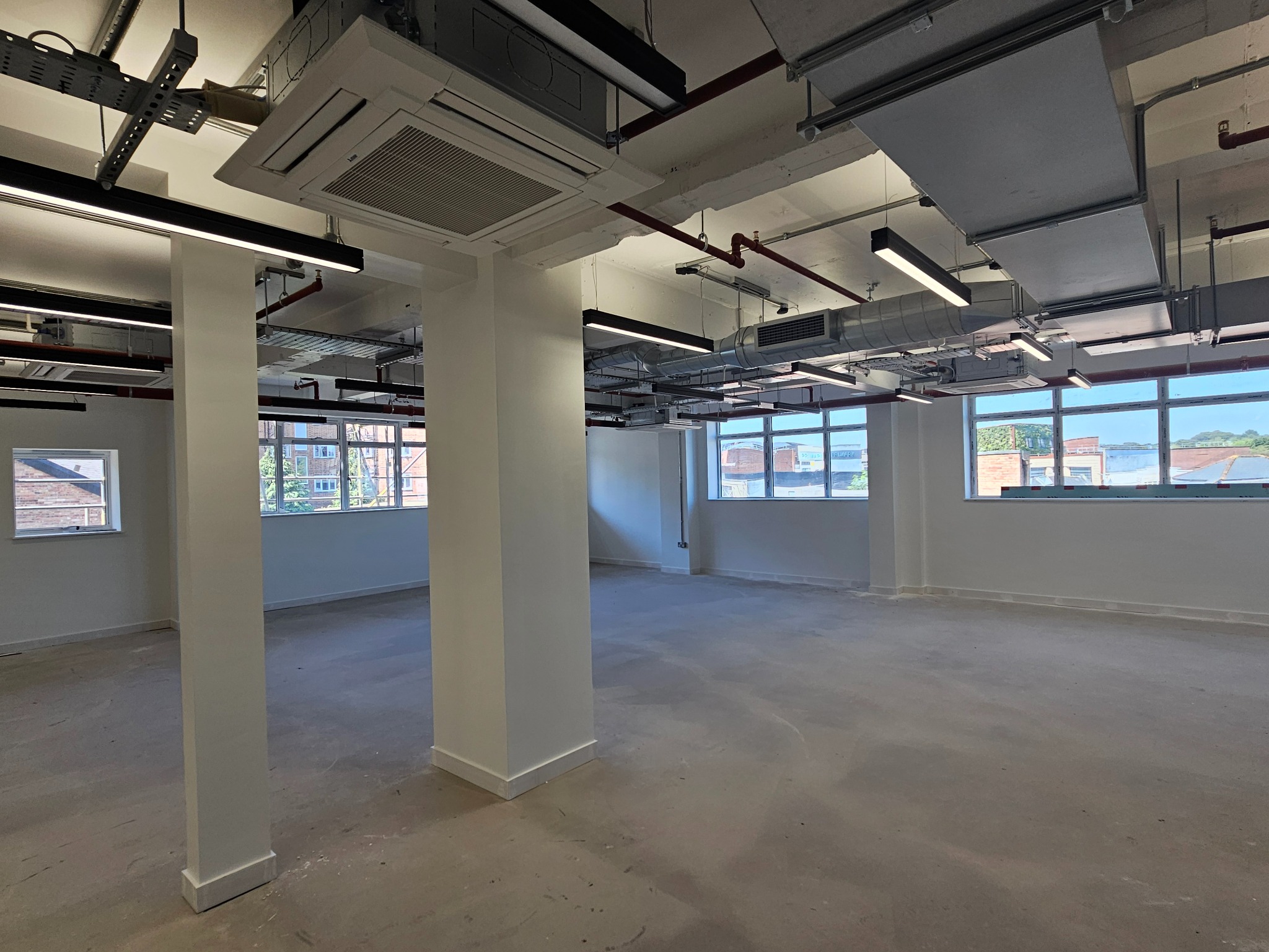Puste, jasne wnętrze biurowe z widoczną instalacją wentylacyjną pod sufitem, świeżo pomalowanymi ścianami i surową betonową podłogą przed wykończeniem.