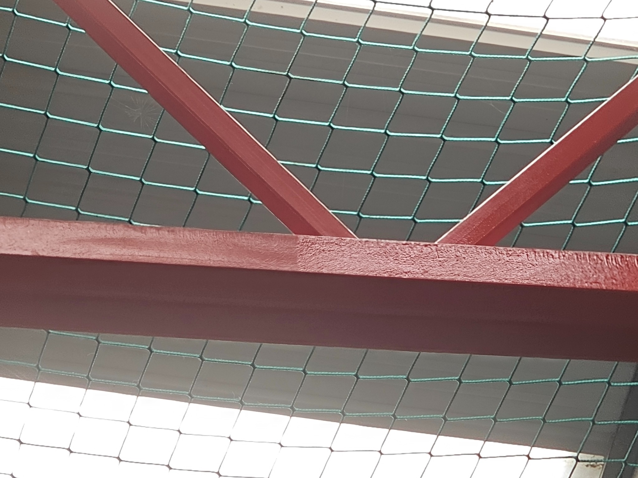 Metalowa konstrukcja pomalowana na czerwono, widoczna od dołu, z siatką zabezpieczającą w tle.