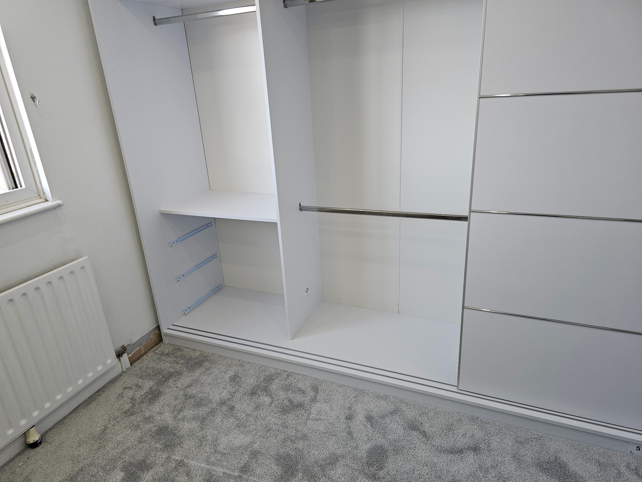 Wnętrze szafy wnękowej z przesuwnymi drzwiami, widoczne półki i drążki na ubrania, szara wykładzina dywanowa na podłodze, grzejnik na ścianie obok.