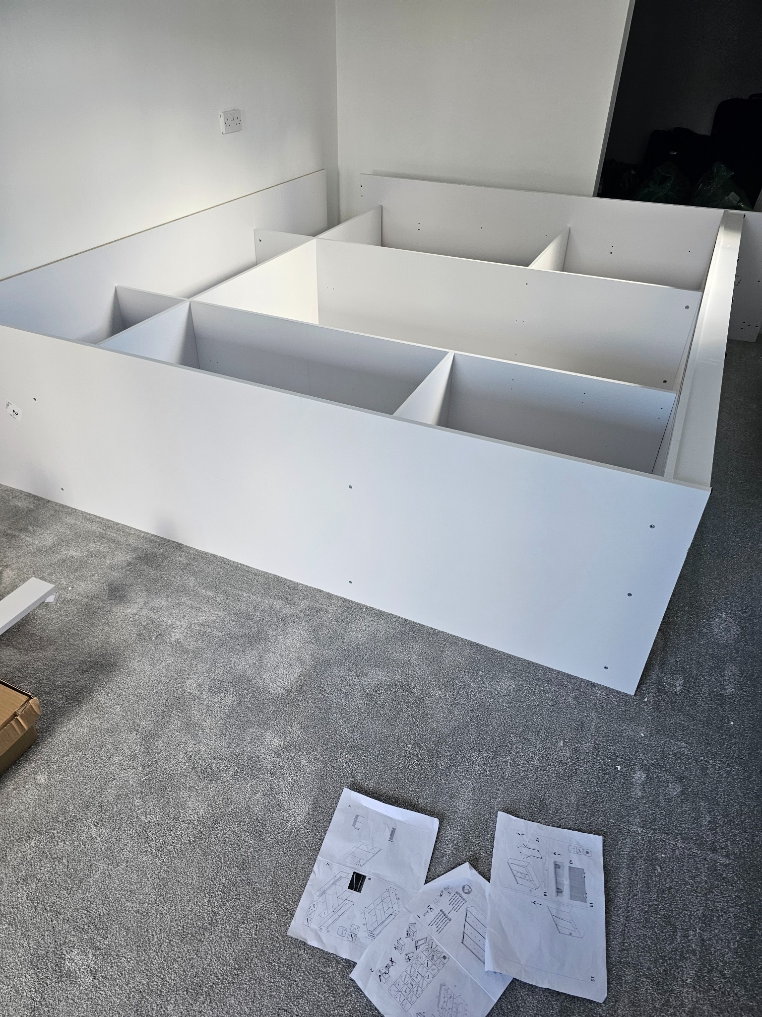 Biała, modułowa konstrukcja regału z podziałem na kwadratowe półki, częściowo zamontowana, z instrukcją montażu rozłożoną na szarym dywanie w tle białej ściany z gniazdkiem elektrycznym.