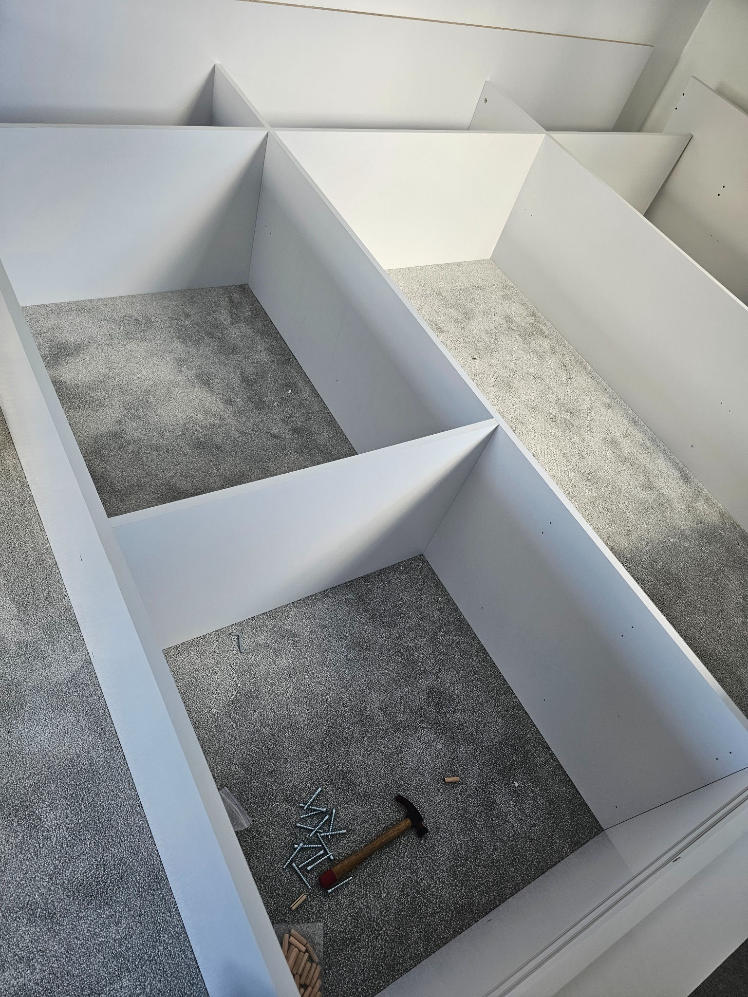Wnętrze szafy wnękowej z białymi półkami, widok z góry na szarą wykładzinę, narzędzia i kołki montażowe leżące na podłodze.
