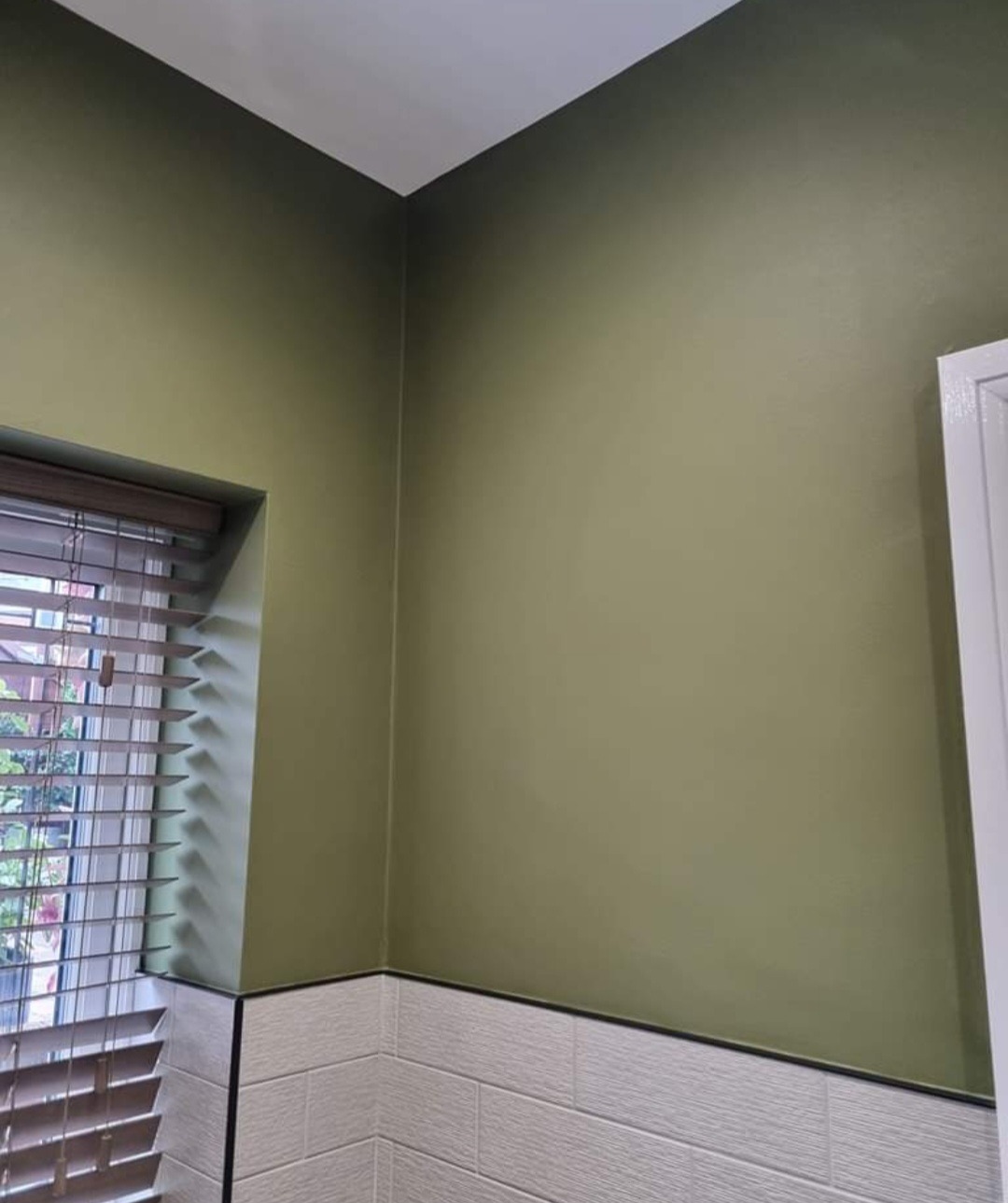 Ściana w odcieniach oliwkowej zieleni, pomalowana na gładko, z widocznym fragmentem okna z żaluzjami i dolną częścią wyłożoną płytkami imitującymi cegłę.