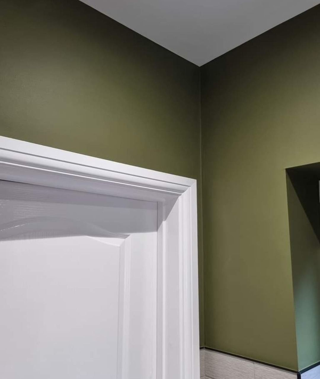 Ściana w odcieniu oliwkowej zieleni, świeżo pomalowana, z widoczną białą, klasyczną ościeżnicą drzwiową i subtelną listwą przypodłogową z szarej mozaiki.