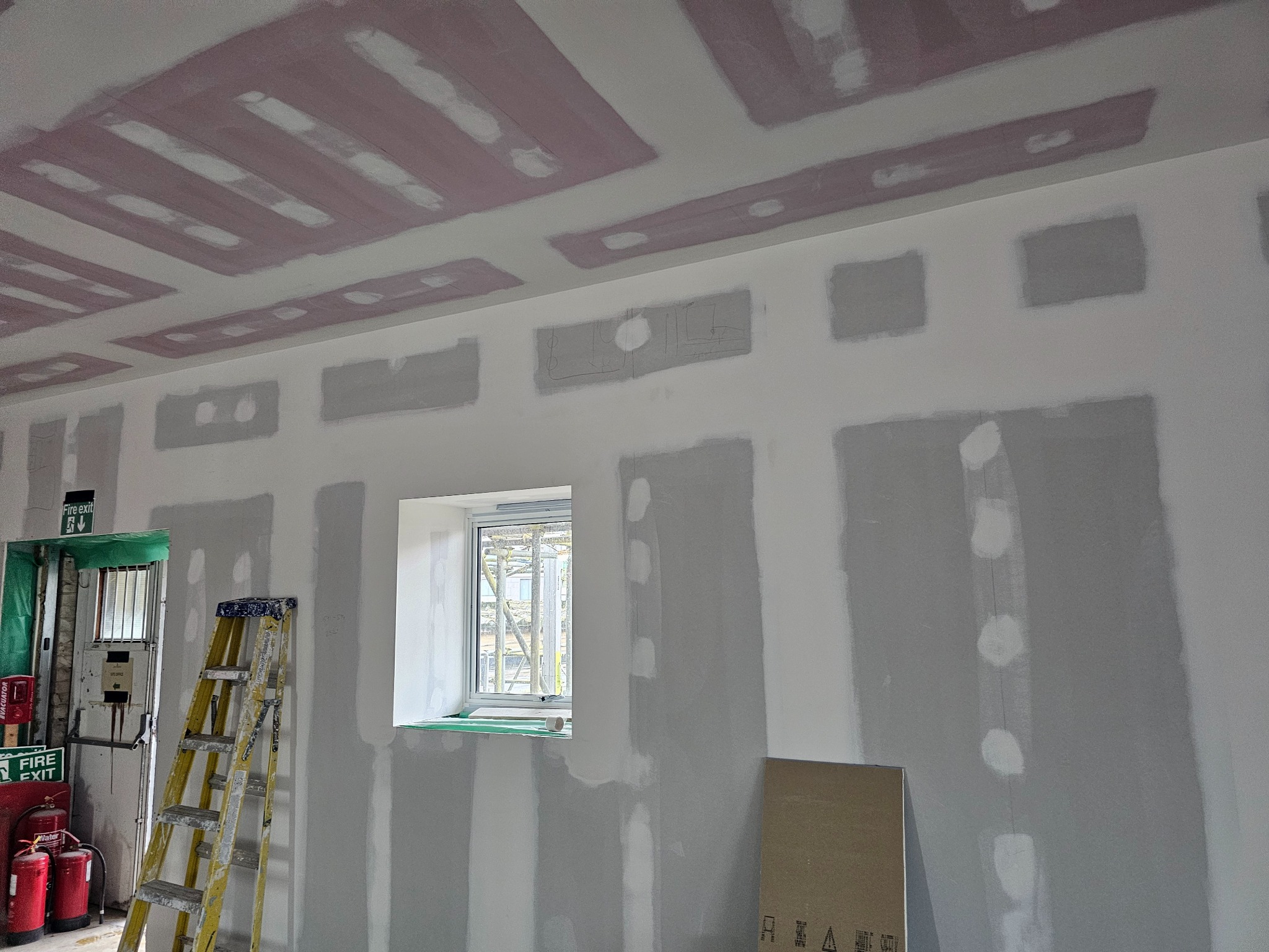 Wnętrze w trakcie prac przygotowawczych: ściany i sufit po szpachlowaniu, widoczna drabina, okno i instalacje przeciwpożarowe.