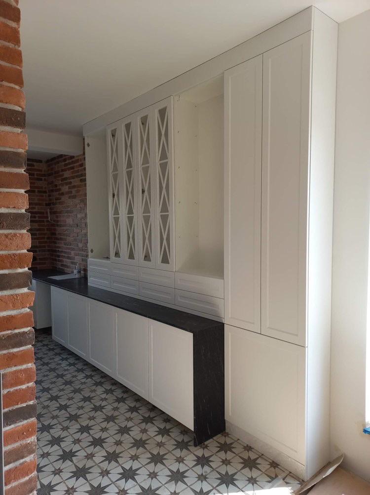 Biała zabudowa kuchenna z czarnym blatem i przeszklonymi szafkami, widok z boku na tle ceglanej ściany i wzorzystej podłogi.