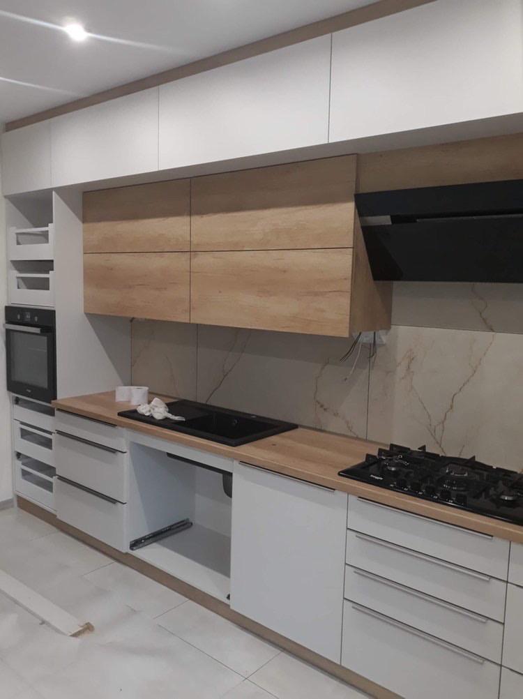 Nowoczesna kuchnia z białymi szafkami dolnymi i górnymi w kolorze drewna, czarny okap i zlewozmywak, marmurowe płytki na ścianie, widoczne niedociągnięcia montażowe