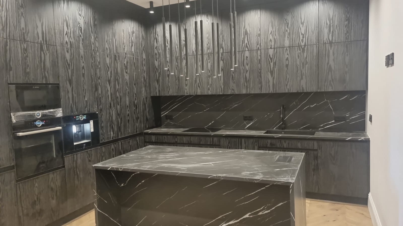 Nowoczesna kuchnia z czarnymi drewnianymi szafkami o wyraźnym usłojeniu, blatem z czarnego marmuru z białymi żyłami, wyspą kuchenną i podłużnymi lampami wiszącymi