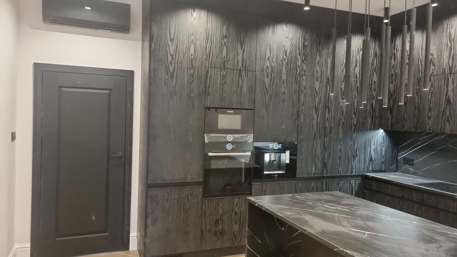 Nowoczesna kuchnia w odcieniach szarości i czerni z drewnianymi frontami szafek, marmurowym blatem wyspy, zabudowanym sprzętem AGD marki Bosch i wiszącymi lampami tubowymi.