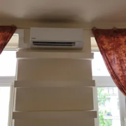 Biały klimatyzator Panasonic zamontowany na ścianie nad oknem, z dekoracyjnymi, pomarańczowo-brązowymi zasłonami po bokach okna.