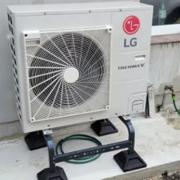 Biała jednostka zewnętrzna pompy ciepła LG Therma V zamontowana na czarnej, metalowej podstawie, widoczna na tle ściany domu z oknem i kranem z podłączonym wężem ogrodowym.