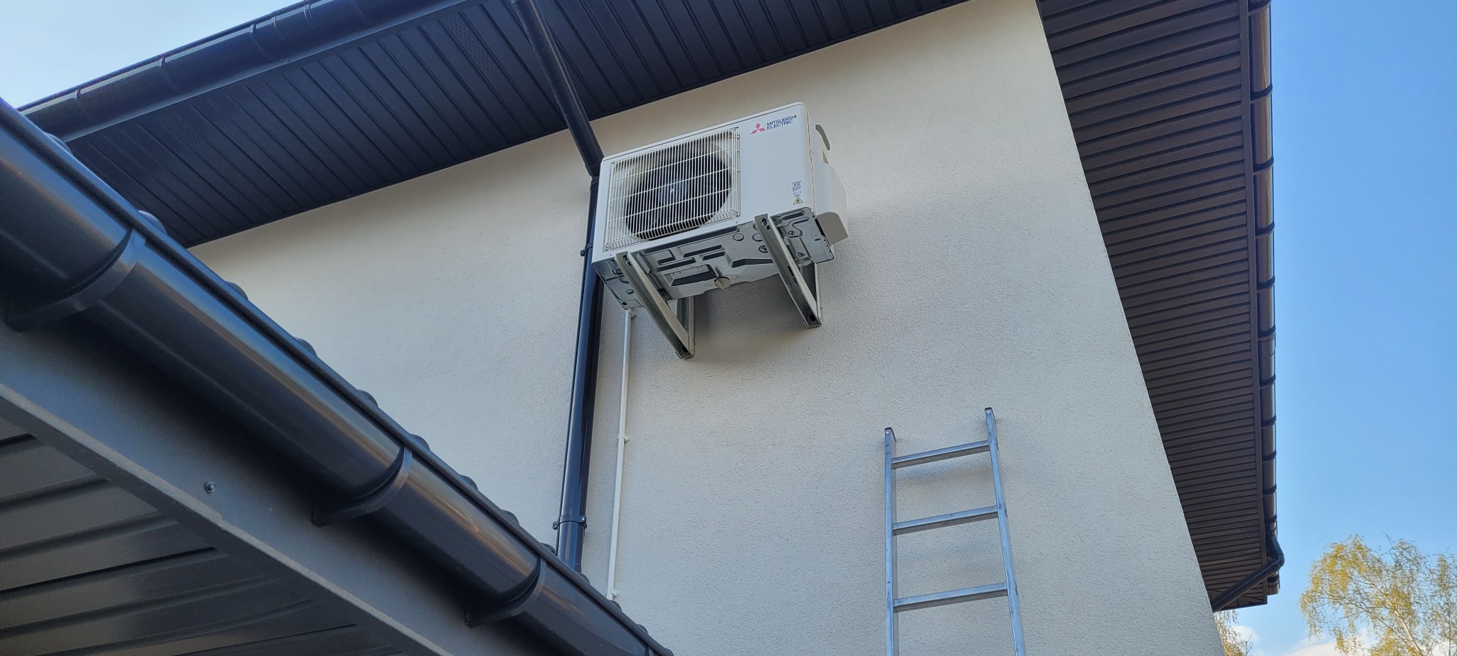 Biała jednostka zewnętrzna klimatyzacji Mitsubishi zamontowana na wspornikach na elewacji budynku, obok rynny spustowej i drabiny aluminiowej.