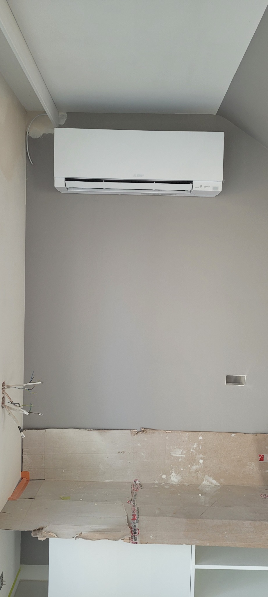 Biały klimatyzator ścienny zamontowany na szarej ścianie w trakcie remontu, widoczne wystające kable i zabezpieczona kartonem dolna część ściany.