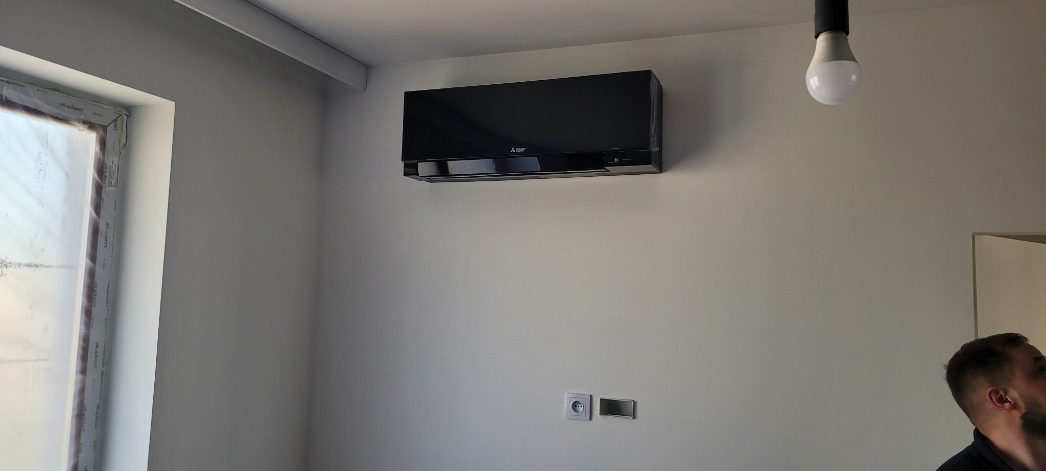 Czarny klimatyzator Mitsubishi Electric zamontowany na białej ścianie w nowym pomieszczeniu, widoczne gniazdka elektryczne i fragment okna z folią ochronną.