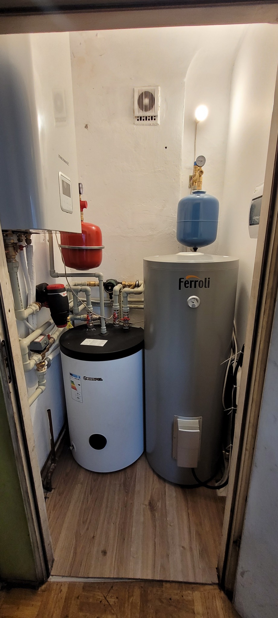 Kompaktowa kotłownia w małym pomieszczeniu: biały zasobnik ciepłej wody użytkowej Kospel obok szarego zasobnika Ferroli, instalacja z rur PCV, reduktor ciśnienia, niska wentylacja ścienna.