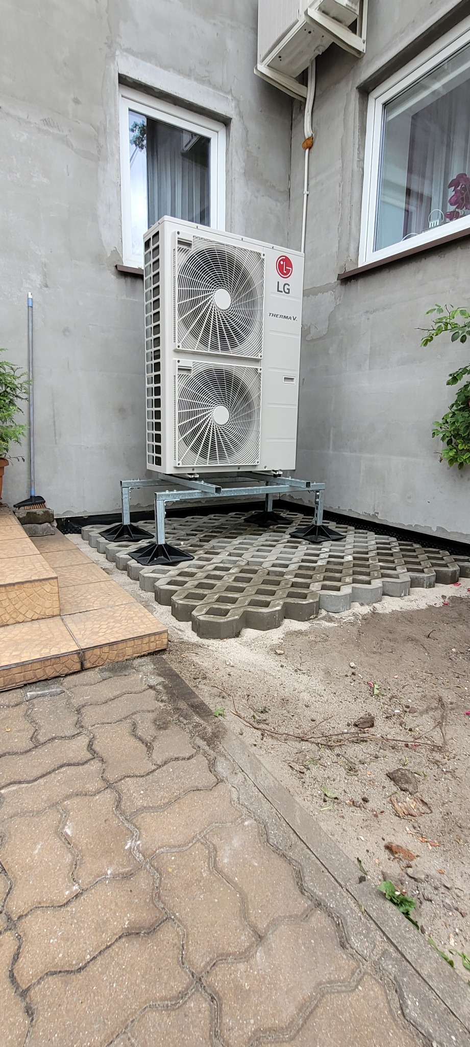 Zewnętrzna jednostka pompy ciepła LG Therma V, zamontowana na metalowej konstrukcji na betonowej ażurowej kostce, przy szarej ścianie budynku z dwoma oknami.