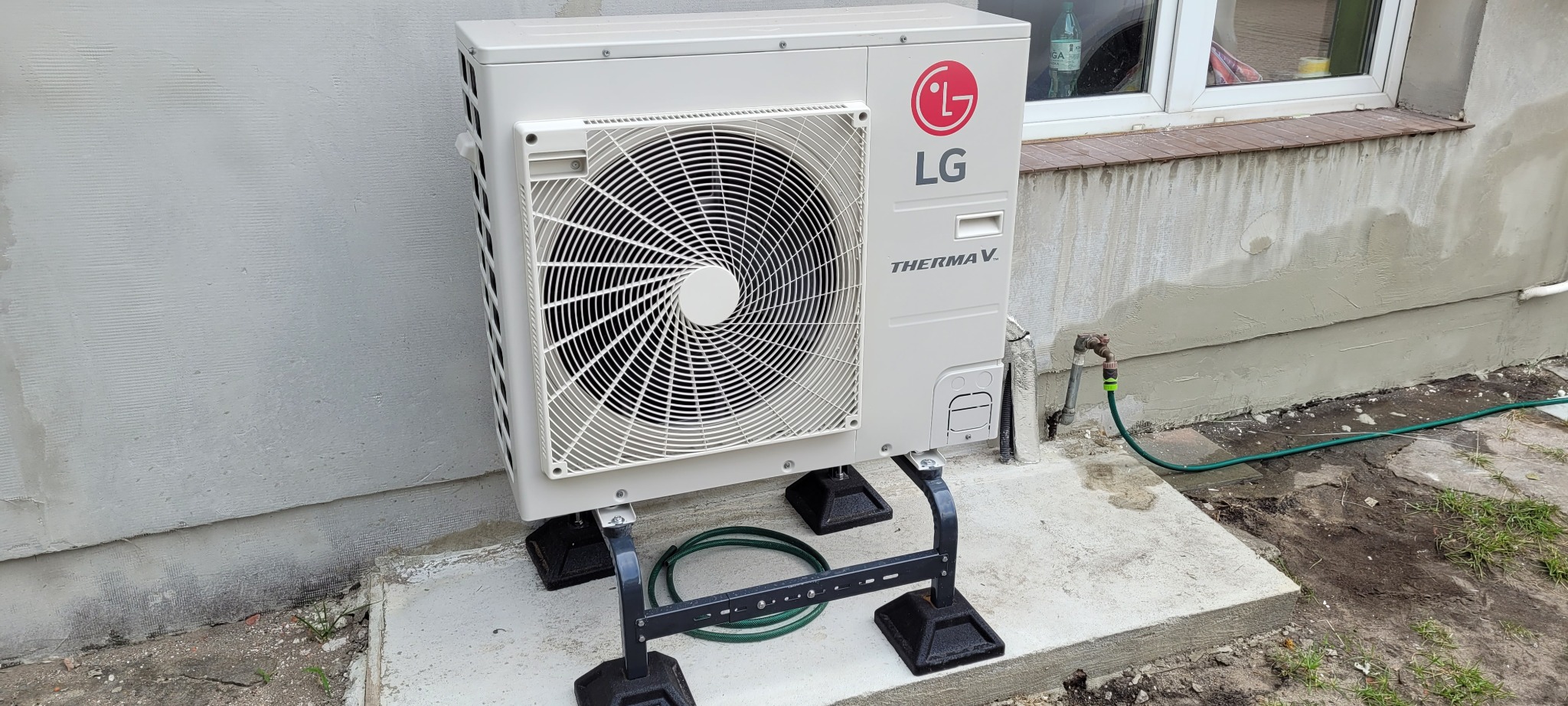 Biała jednostka zewnętrzna pompy ciepła LG Therma V zamontowana na czarnej, metalowej podstawie, widoczna na tle ściany domu z oknem i kranem z podłączonym wężem ogrodowym.