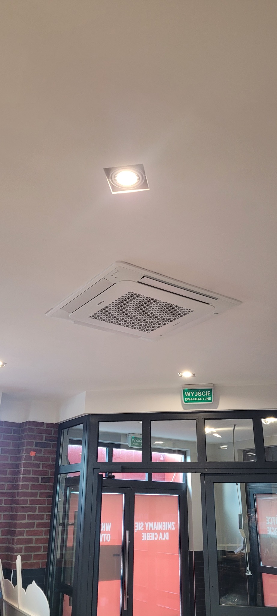 Biała, sufitowa klimatyzacja kasetonowa Samsung zamontowana w pomieszczeniu komercyjnym, widoczna lampa sufitowa i znak ewakuacyjny.