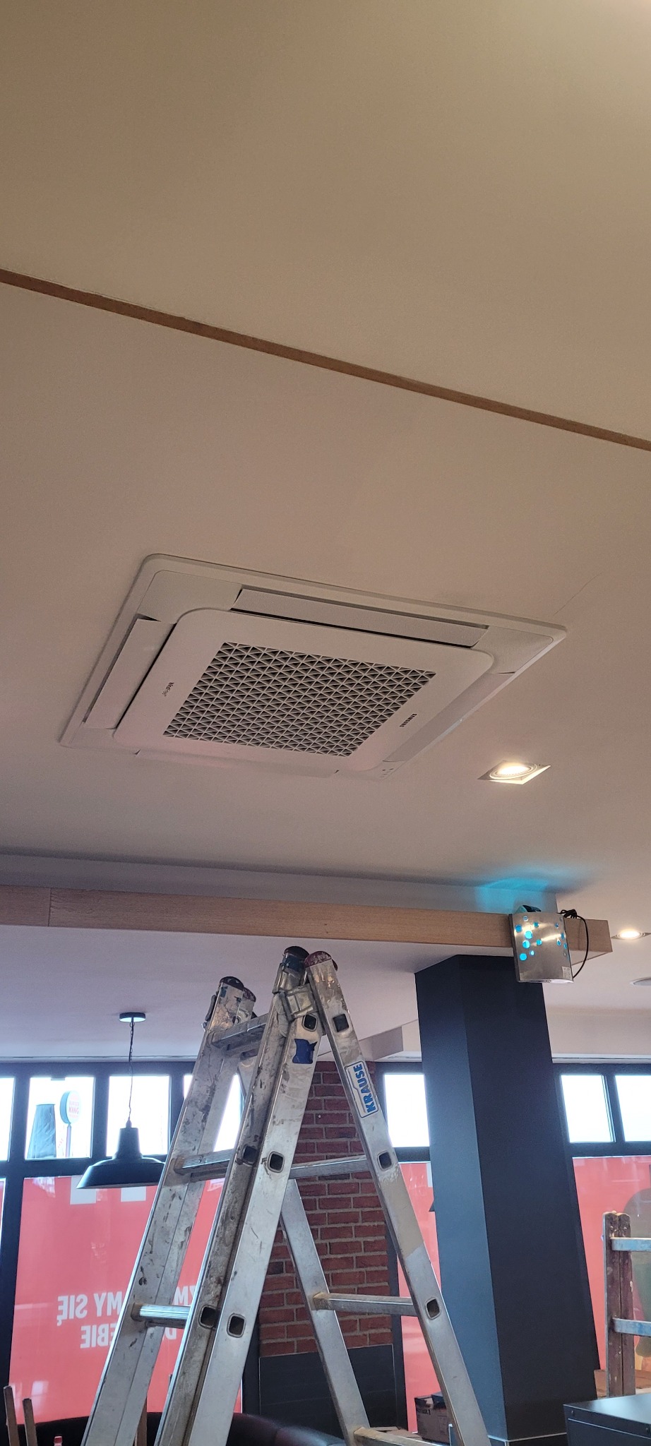 Kasetonowa jednostka klimatyzacji Samsung zamontowana w suficie lokalu, widoczna z dołu z perspektywy drabiny Krause. W tle widoczne elementy wystroju wnętrza, w tym lampa i ceglana ściana.