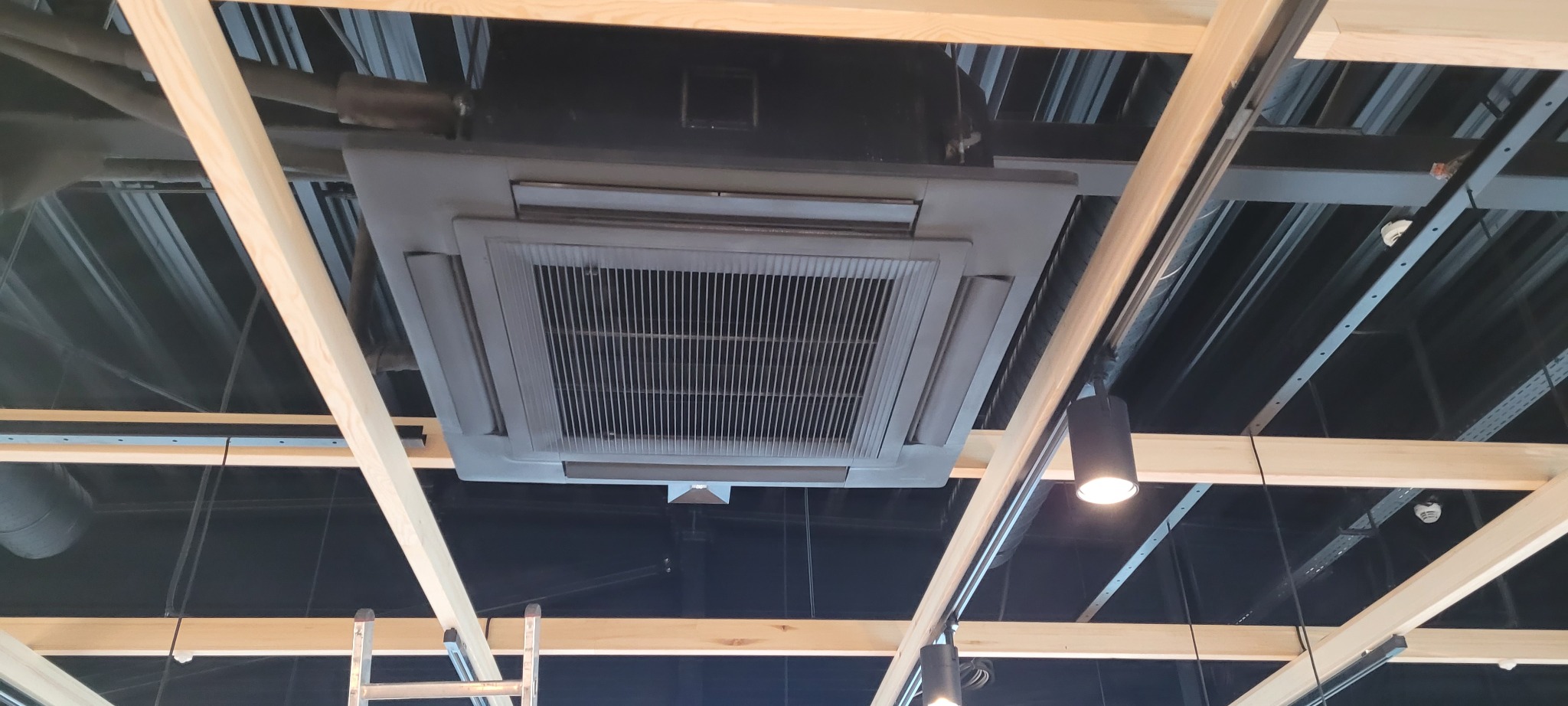 Widok z dołu na zainstalowaną kasetonową jednostkę klimatyzacji w pomieszczeniu komercyjnym, widoczne elementy konstrukcji sufitu podwieszanego i oświetlenie punktowe.