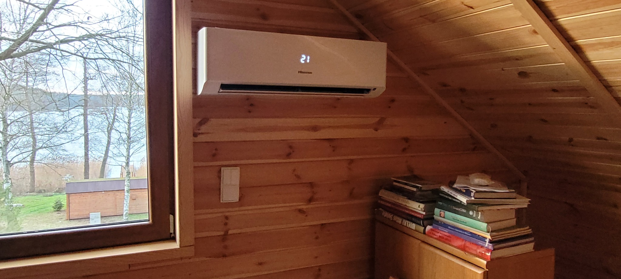 Klimatyzator Hisense zamontowany na drewnianej ścianie pod skosem, widok przez okno na jezioro i drzewa, obok stos książek.