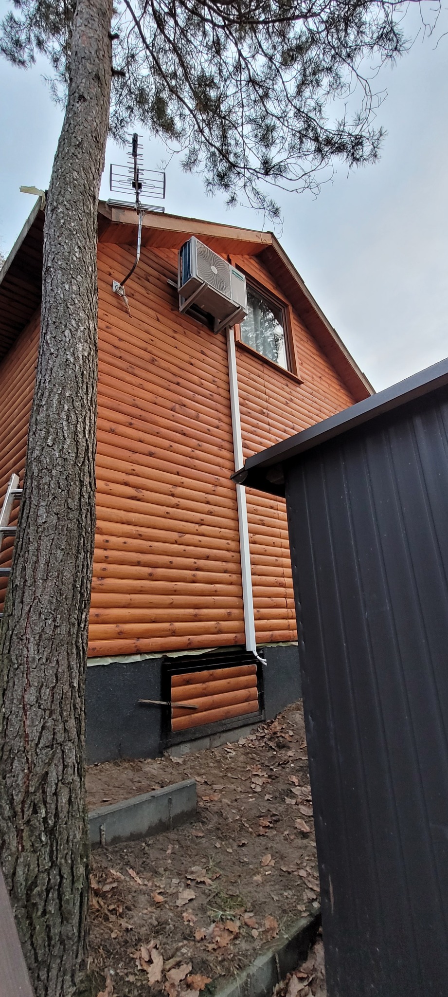 Zewnętrzna jednostka klimatyzacji zamontowana na drewnianej ścianie domu obok okna i anteny telewizyjnej, widok z dołu.