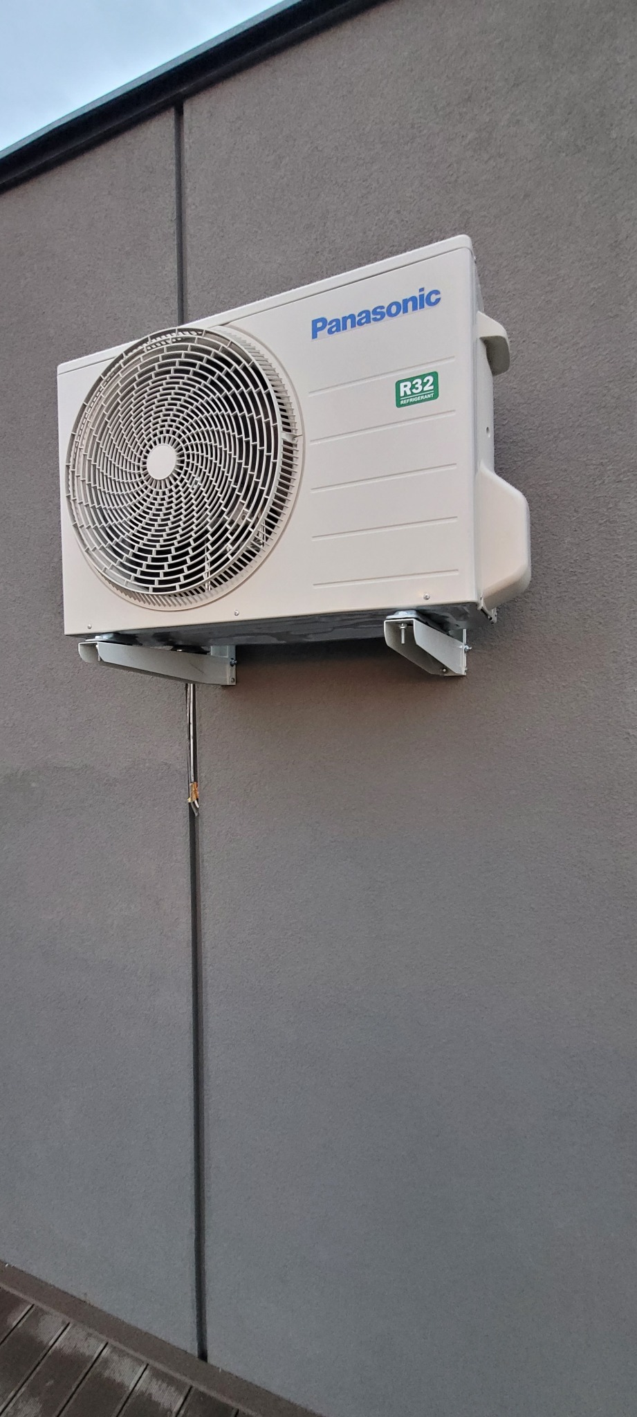 Zewnętrzna jednostka klimatyzacji Panasonic zamontowana na szarej ścianie budynku, widoczne metalowe wsporniki i rurka odprowadzająca skropliny.