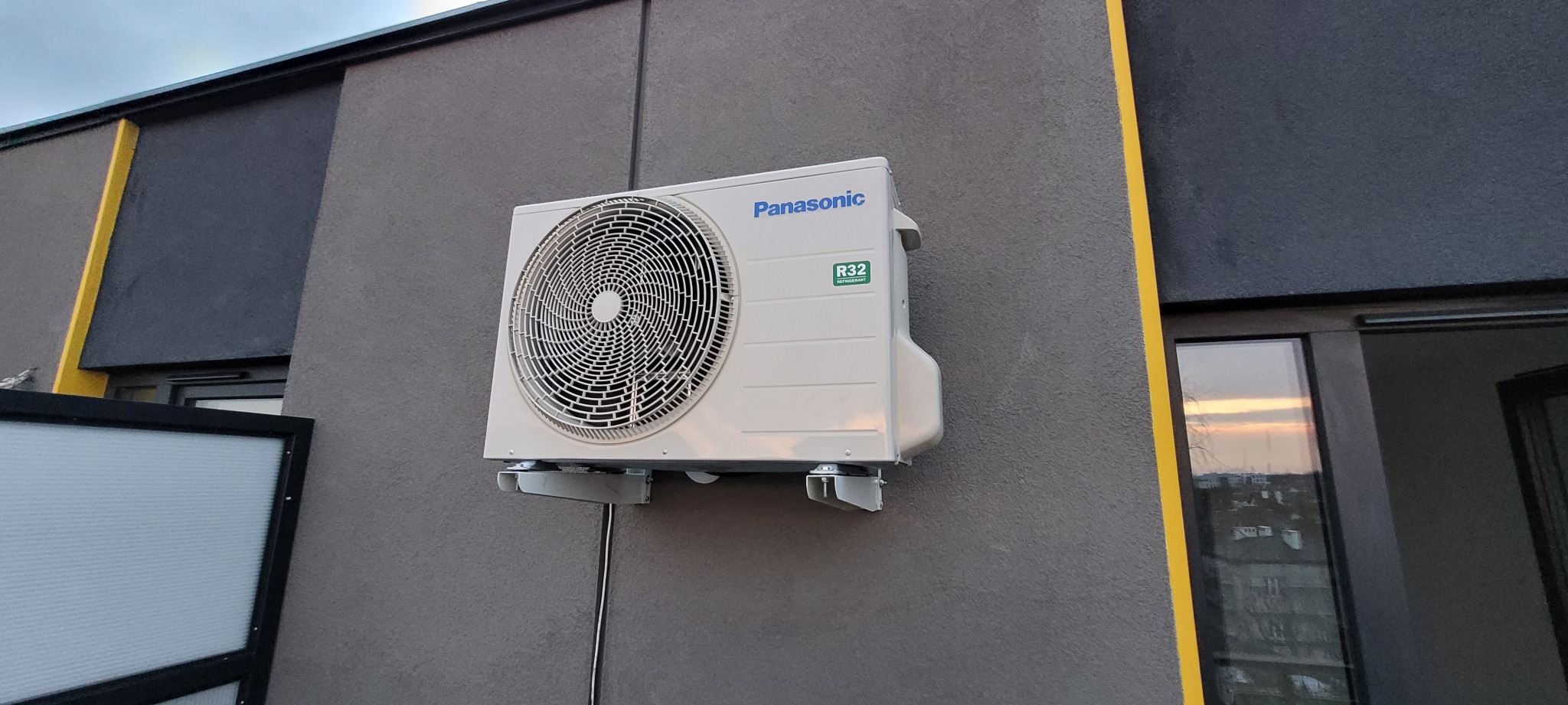Zewnętrzna jednostka klimatyzacji Panasonic zamontowana na szarej ścianie budynku z widocznym przewodem i oknem odbijającym zachód słońca.