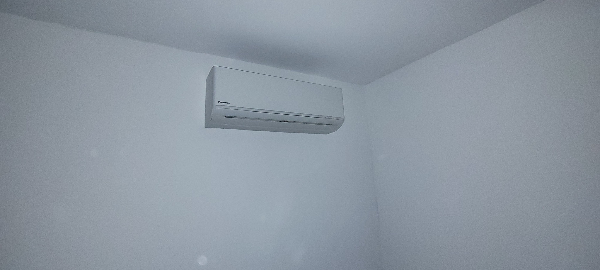 Biała jednostka klimatyzacyjna Panasonic zamontowana na białej ścianie w rogu pokoju, widok z dołu.