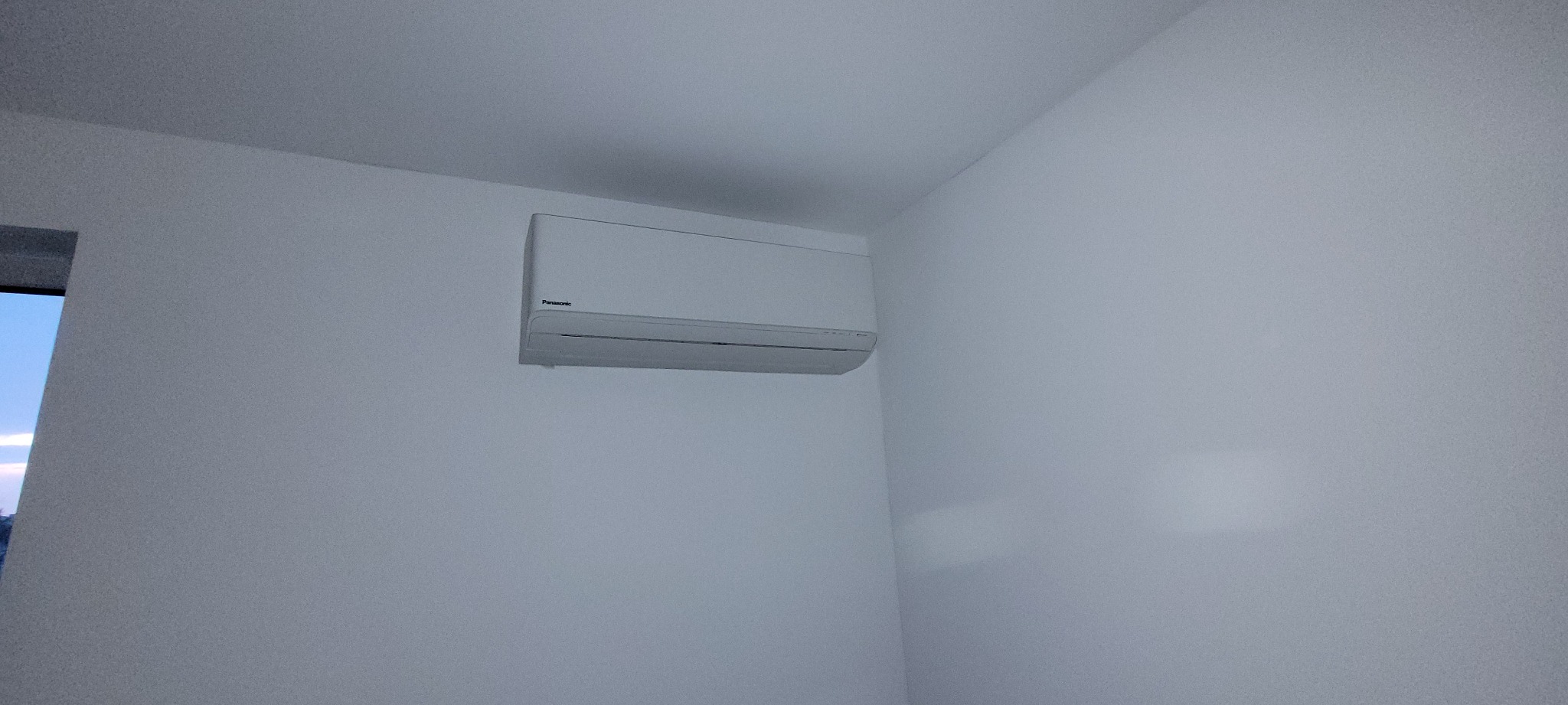 Zamontowana, biała klimatyzacja Panasonic na białej ścianie w minimalistycznym wnętrzu, widoczny fragment okna po lewej stronie.