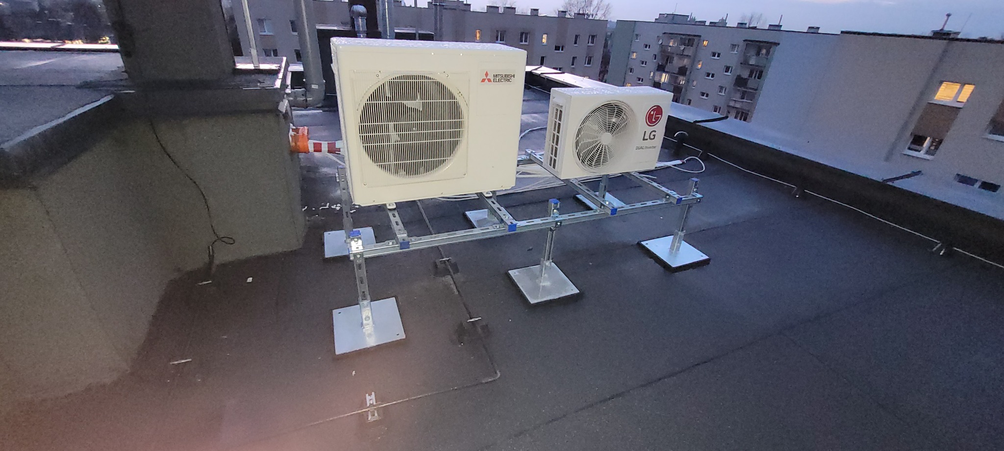 Dwa klimatyzatory (Mitsubishi Electric i LG Dual Inverter) zamontowane na metalowej konstrukcji na dachu budynku wielorodzinnego, widoczne bloki mieszkalne w tle.