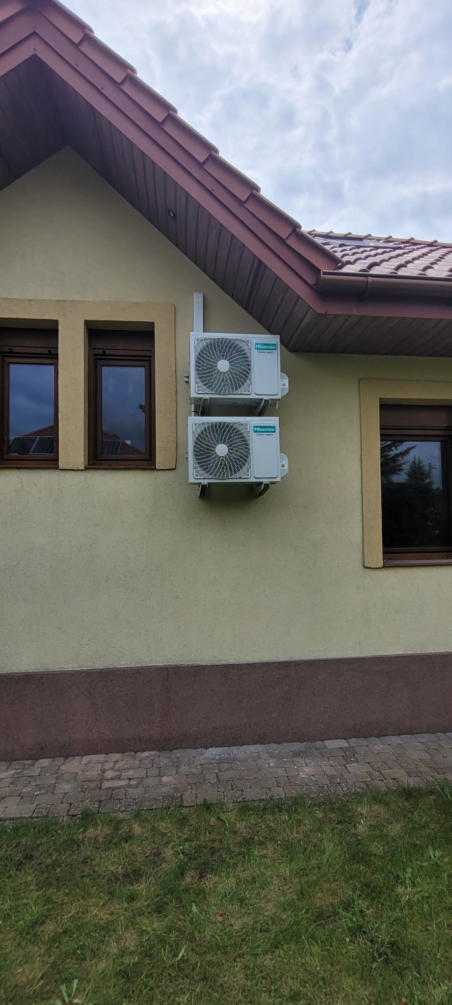 Dwa białe klimatyzatory marki Hisense zamontowane jeden nad drugim na elewacji domu z brązowymi oknami i dachem z dachówki ceramicznej. Widoczny trawnik i fragment chodnika z kostki.