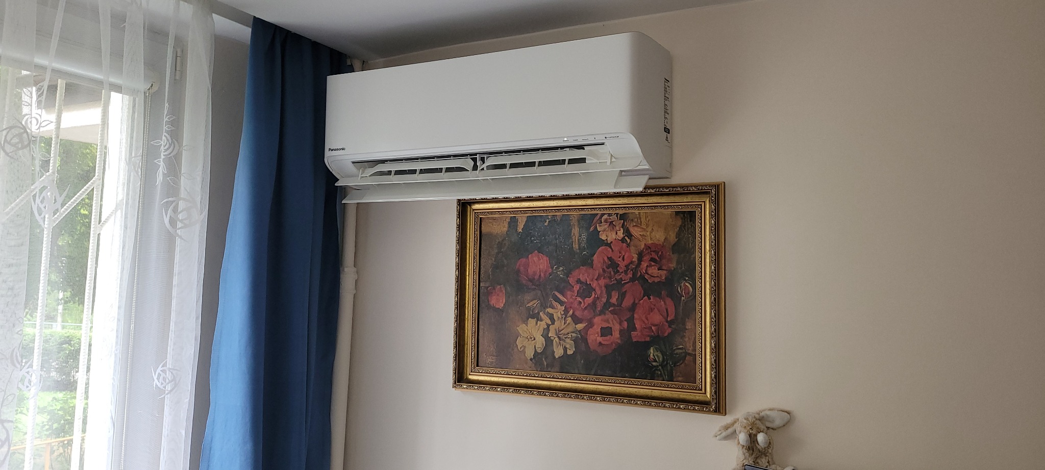 Biały klimatyzator Panasonic zamontowany nad obrazem w złotej ramie, obok okna z niebieskimi zasłonami.