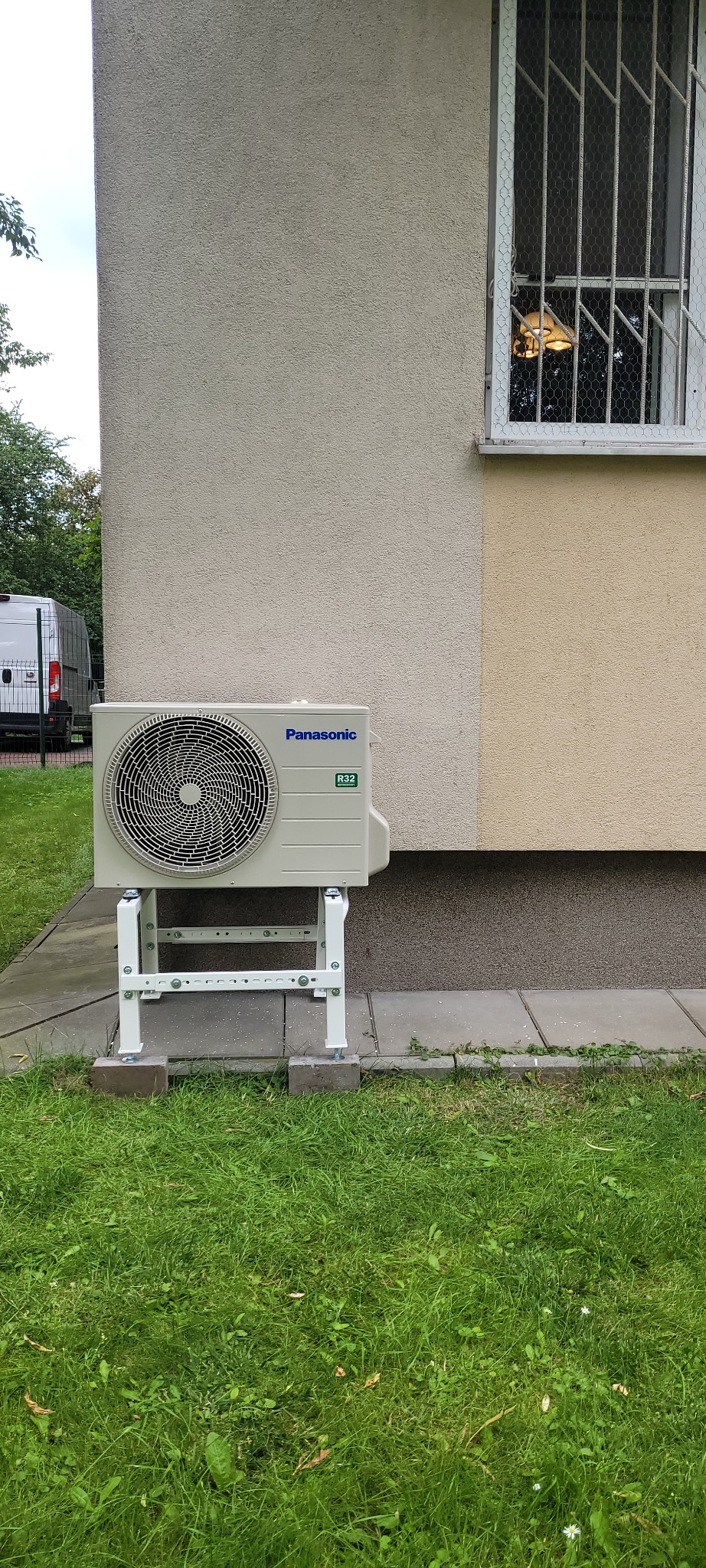 Zewnętrzna jednostka klimatyzacji Panasonic na białym stelażu, zamontowana przy ścianie budynku w Leoncinie. W tle widoczne okno z kratami i zieleń.