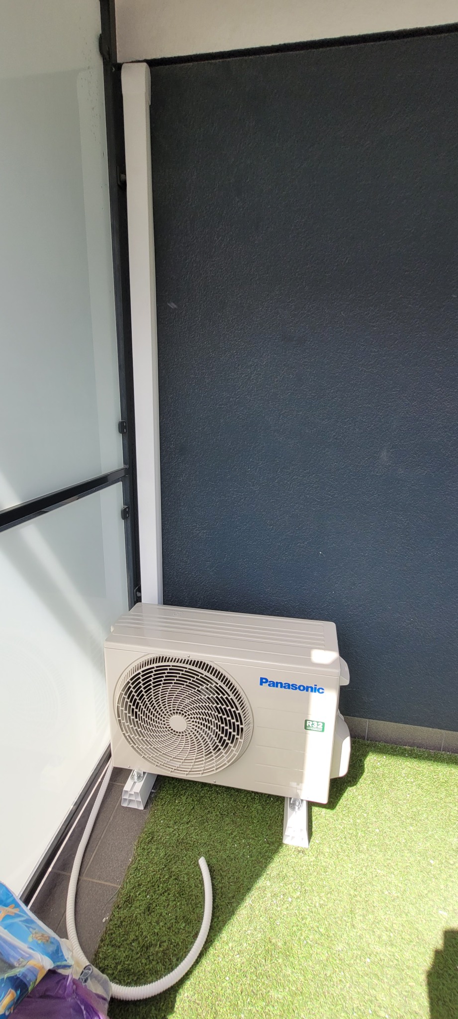 Zewnętrzna jednostka klimatyzacji Panasonic zamontowana na balkonie wyłożonym sztuczną trawą, widoczna rurka odprowadzająca skropliny.