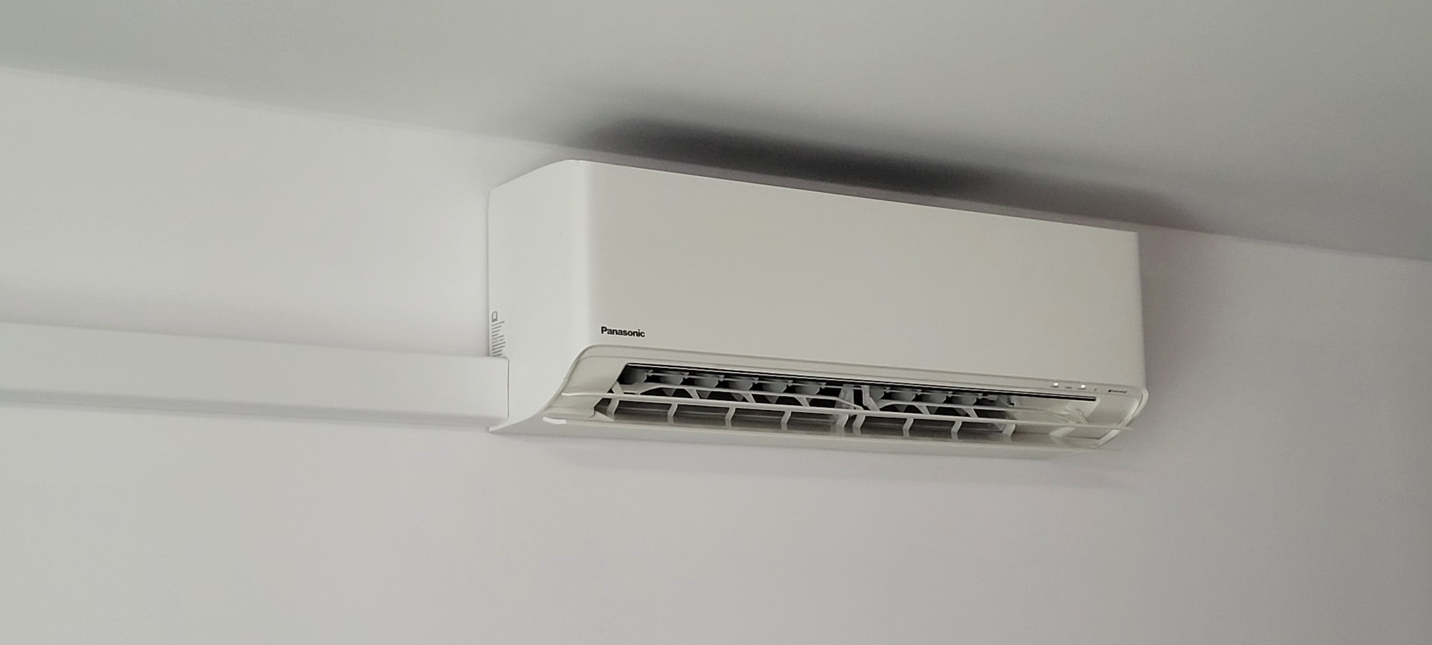 Biała klimatyzacja Panasonic zamontowana na ścianie w jasnym pomieszczeniu, widoczne diody sygnalizacyjne.
