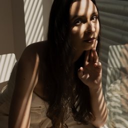 Ewa Kalinowska - Portret kobiety z długimi ciemnymi włosami, siedzącej w fotelu, oświetlonej przez promienie słoneczne padające przez żaluzje, z ręką podpartą pod brodą i delikatnym uśmiechem.