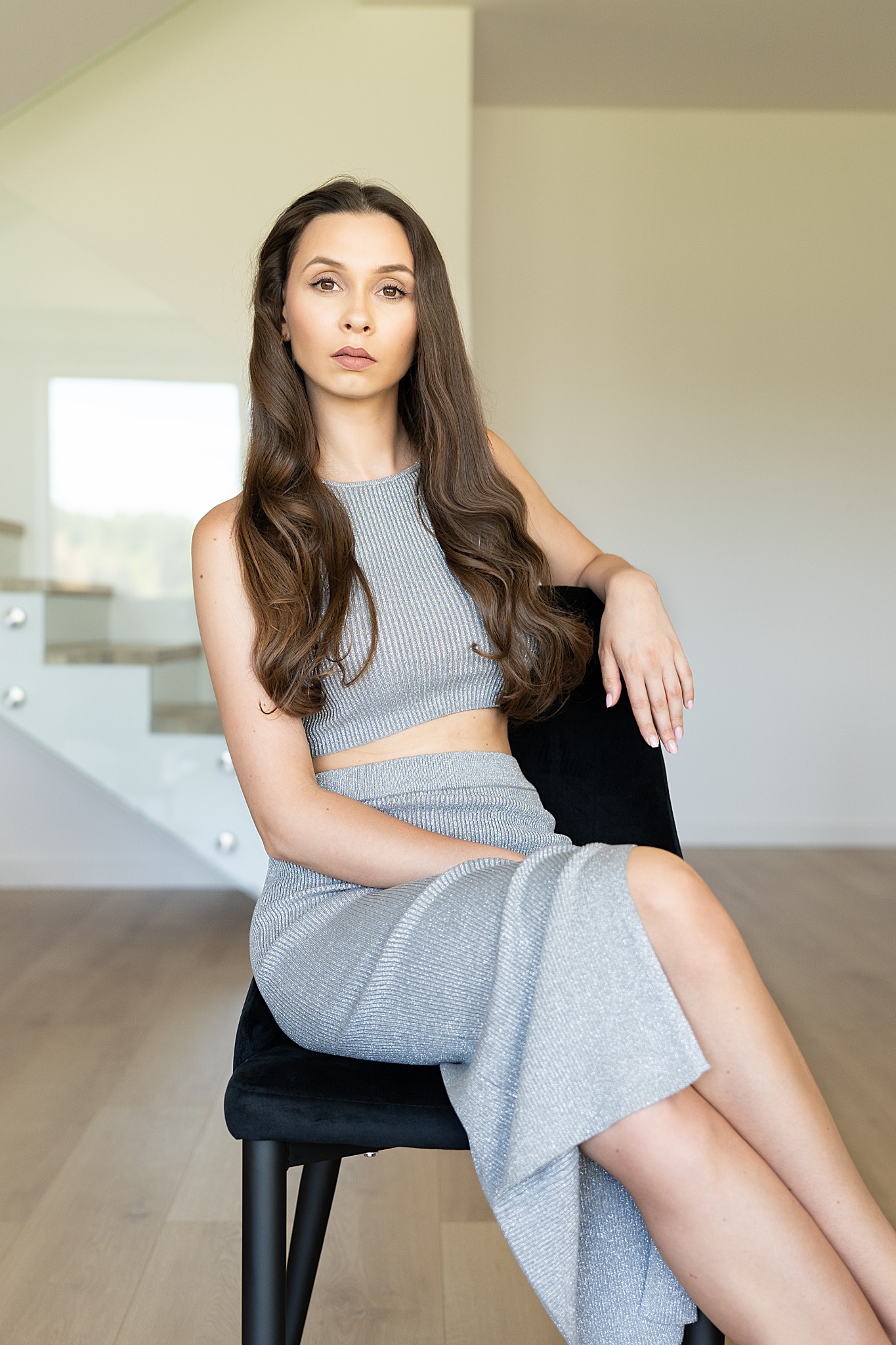 Portret kobiety w srebrnym, prążkowanym komplecie siedzącej na czarnym krześle na tle minimalistycznego wnętrza z białymi schodami w tle.
