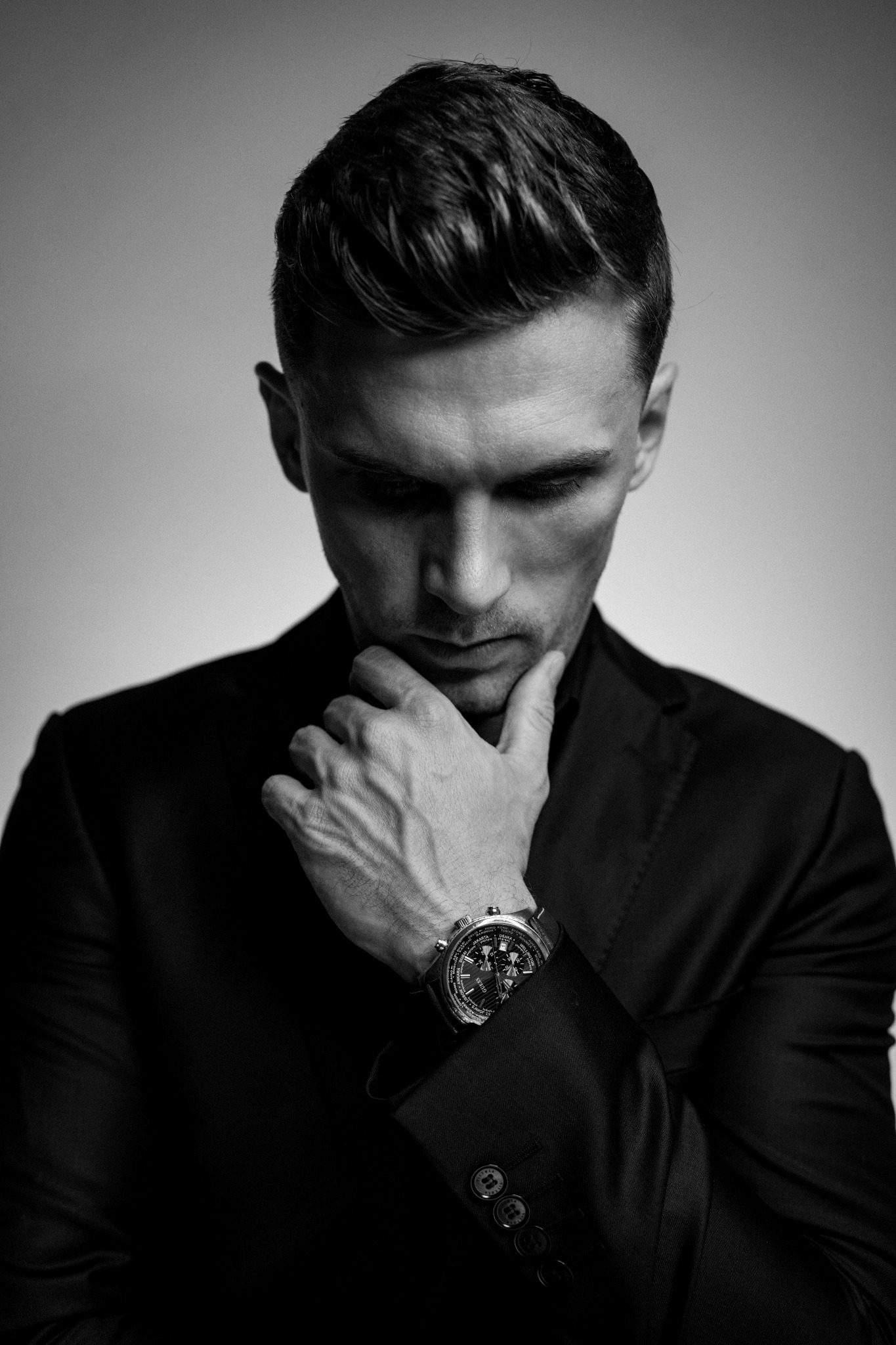 Czarno-białe zbliżenie na zamyślonego mężczyznę w garniturze, dotykającego brody, z widocznym zegarkiem na nadgarstku i starannie ułożoną fryzurą.